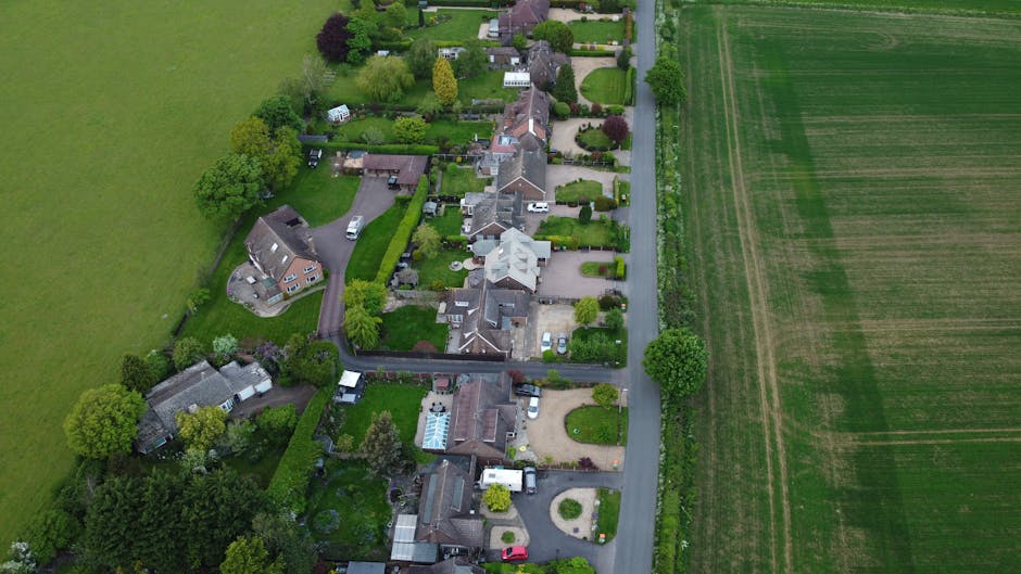 An aerial view of a suburban residential area showing a row of houses with neatly maintained gardens along a quiet street. The gardens feature a variety of lush green trees, well-trimmed hedges, and vibrant flower beds, with some lawns exhibiting freshly mown grass. Several driveways and pathways are visible, leading to garages and entrances, with garden tools like lawn mowers and garden shears placed nearby, indicating ongoing garden maintenance. The surrounding landscape includes expansive green fields and open spaces, with visible textures of soil, grass, and leaves, all under soft daylight. This image highlights the well-kept outdoor spaces typical of residential gardens in a countryside setting, with subtle signs of landscaping work, such as leaf debris and neatly edged borders, reflecting professional garden care by Gardeners Watford.