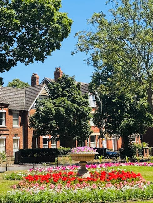 Front garden of a residential property featuring a large pink flowering cherry blossom tree in full bloom, with delicate clusters of pink flowers covering its branches. The garden has a well-maintained green hedge forming a border along a low brick wall, with a paved pathway leading to a dark green front door situated under a white porch with two white columns. The house exterior is painted in a light pastel color, and the overall scene is illuminated by natural daylight, suggesting mild weather conditions suitable for garden maintenance. Gardeners Watford may focus on periodic hedge trimming and pruning of the cherry blossom to preserve its shape and health, contributing to the landscaping work for this front garden.