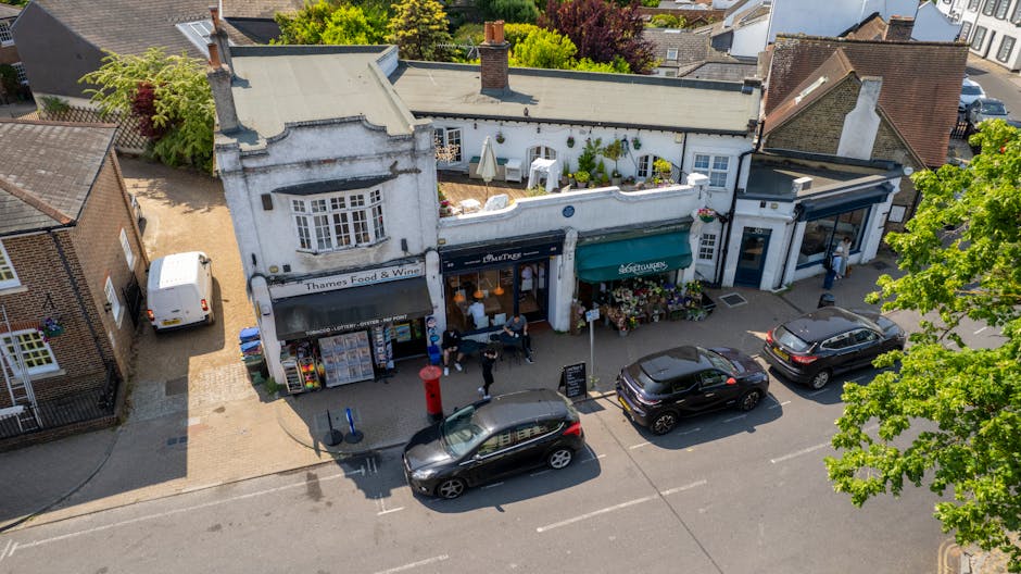 An aerial view of a quaint high street featuring a small row of white-painted shops with black and green awnings, including a Thames Food & Wine store; outdoor seating with white umbrellas, and people walking along the paved sidewalk. In front of the shops, three cars are parked parallel to the curb, one of which is a black SUV. To the left, a narrow alley leads to the back of the buildings, with a white van parked nearby. Surrounding the scene are residential houses with brick facades and tiled roofs, and lush green trees with fresh leaves casting shadows on the street below. The lighting suggests a bright, sunny day, illuminating the textures of the buildings, foliage, and vehicles. This street scene highlights typical urban landscaping and outdoor activity in Watford, captured in a natural and detailed manner.